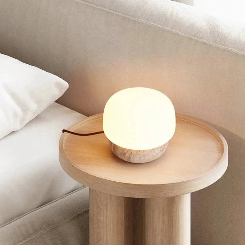 A small, glowing table lamp with a round white glass shade and a textured, light-colored base sits on a round light wooden side table. The lamp's brown cord extends to the left. The table is positioned next to a bed with white linens and a headboard.