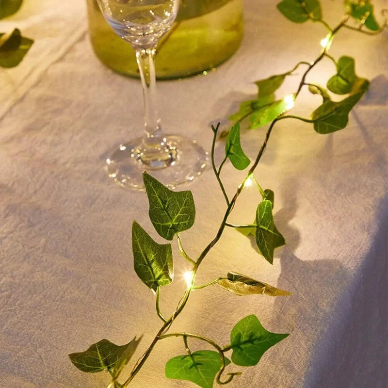A string of warm white fairy lights intertwined with artificial green ivy leaves lies on a white tablecloth. Part of a glass and a blurred container are visible in the background.