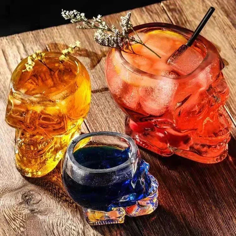 Three different sized glass skull cups filled with a reddish-orange cocktail, a small blue drink, and a golden drink, on a wooden table.