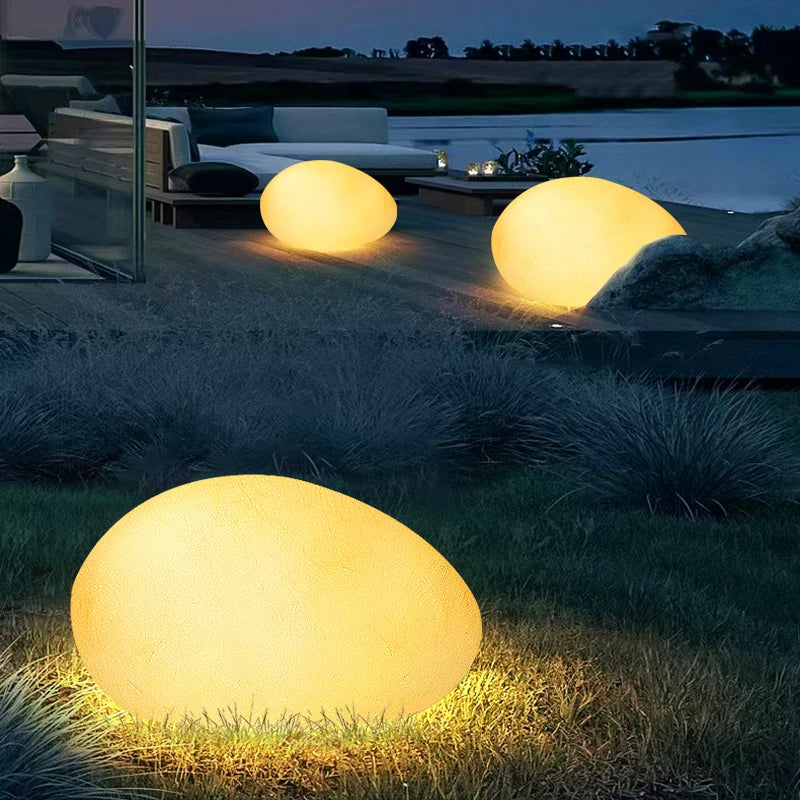 Three illuminated, round egg-shaped lights on a wooden patio with modern outdoor furniture, overlooking a body of water at dusk.