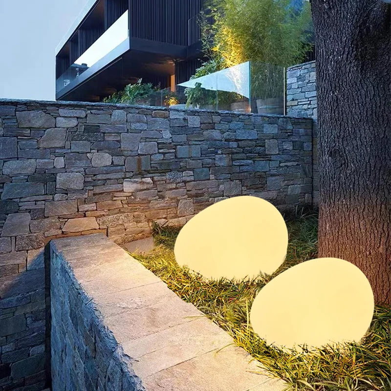 Two illuminated, round egg-shaped lights on a grassy area next to a stone wall and a modern building with a glass balcony.