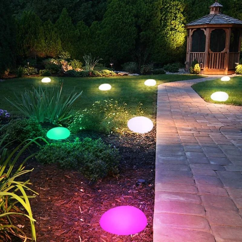 A nighttime view of a garden with a winding brick path illuminated by several glowing, egg-shaped lights. Most of the lights emit a soft white glow, while one near the foreground is pink and another amongst green plants is green. A wooden gazebo is visible in the upper right corner, surrounded by trees and greenery lit by various outdoor lights.