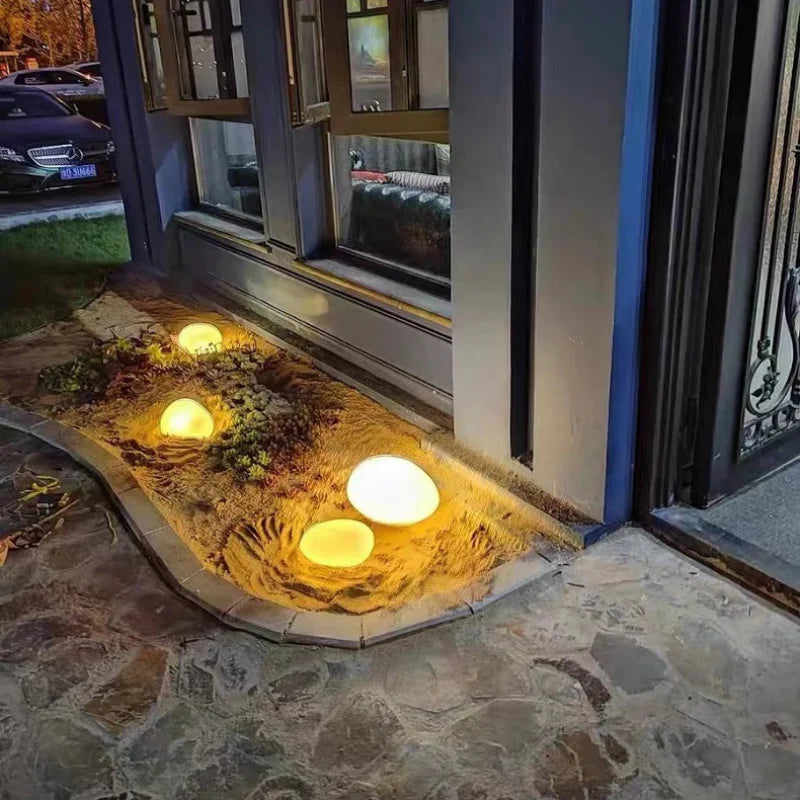 A ground-level view of a narrow garden bed alongside a house, featuring several round, glowing white lights nestled among plants and light-colored mulch. The house has windows and a door visible, and a car is parked in the background. The ground is paved with stone tiles.