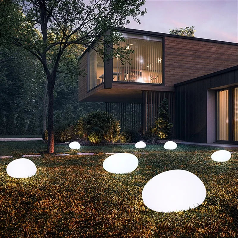 A modern house with a cantilevered section and large windows, surrounded by a yard with glowing egg-shaped lights at dusk.
