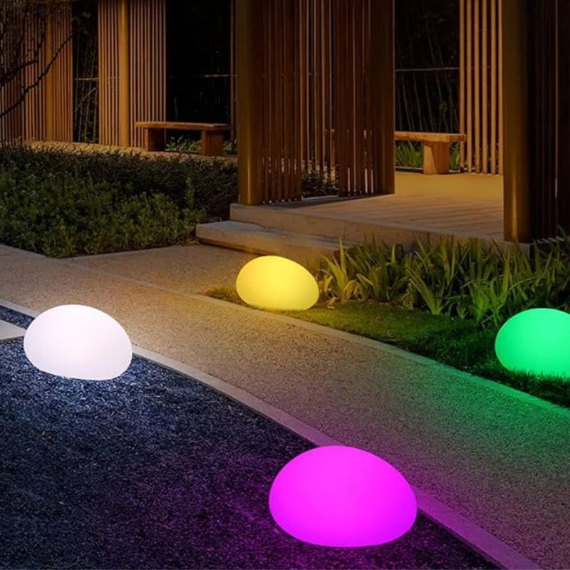 A nighttime shot of a modern outdoor space featuring four glowing, egg-shaped lights in different colors: white, yellow, green, and pink. The lights are placed along a walkway made of small, dark stones, bordered by concrete and patches of green grass and plants. In the background, a building with vertical wooden slats and warm interior lighting can be seen, along with two simple wooden benches.