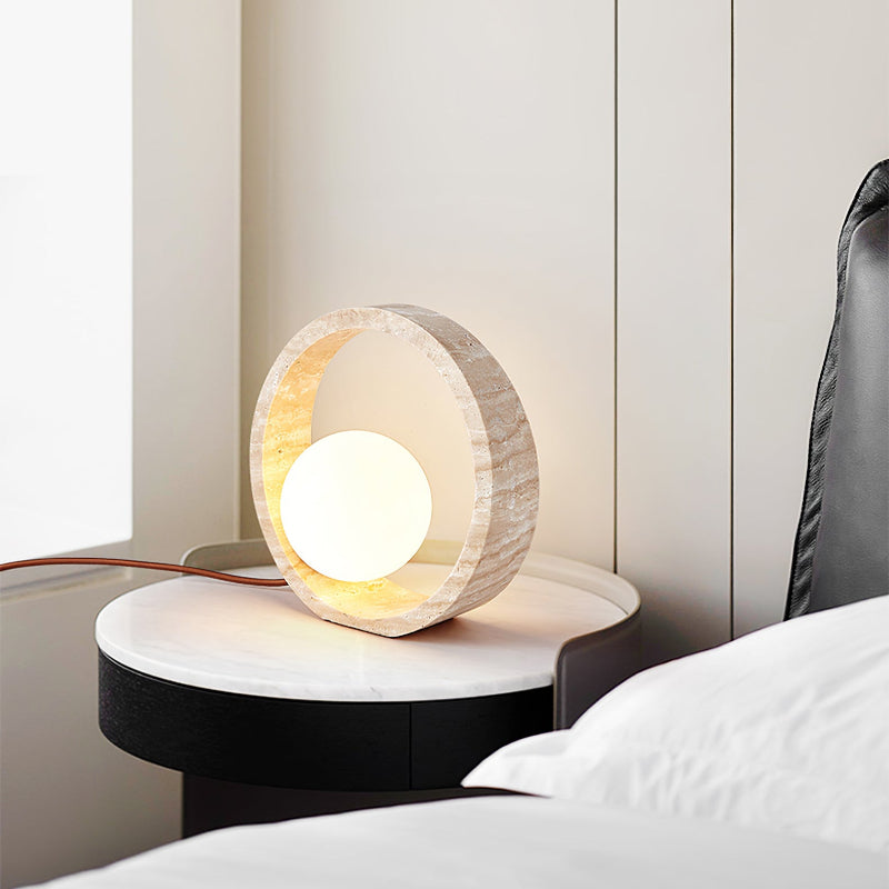 A circular table lamp with a textured, light-colored outer ring and a glowing white spherical inner light sits on a round white and black nightstand. The lamp's brown cord extends to the left towards a window. The nightstand is next to a bed with white linens and a black headboard against a light-colored wall. The inner part of the outer ring emits a warm glow.