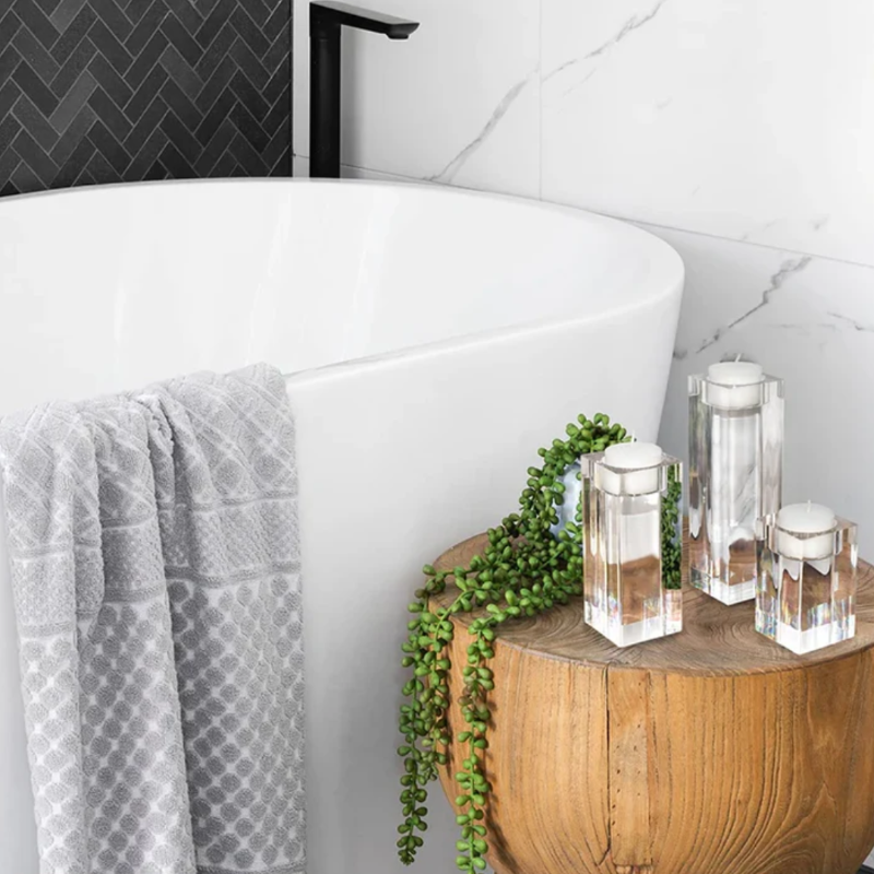 Three clear, multifaceted crystal jars with silver lids sit on a rustic wooden stump table in a bathroom setting. A trailing green plant drapes over the side of the table. A white freestanding bathtub with a grey patterned towel hanging over its edge is visible in the background, along with white marble and dark herringbone tiles on the walls.