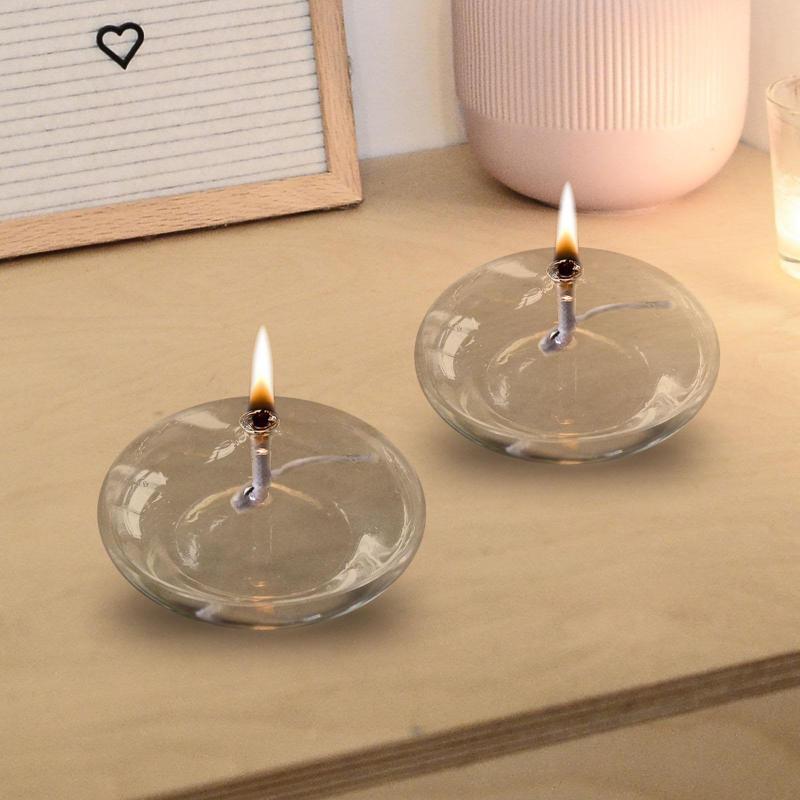 Two small, round glass oil lamps with lit wicks sit on a light brown wooden surface. The flames are bright, and the lamps are partially filled with clear oil. A white letter board with a black heart symbol and a light pink ribbed vase are in the blurred background.