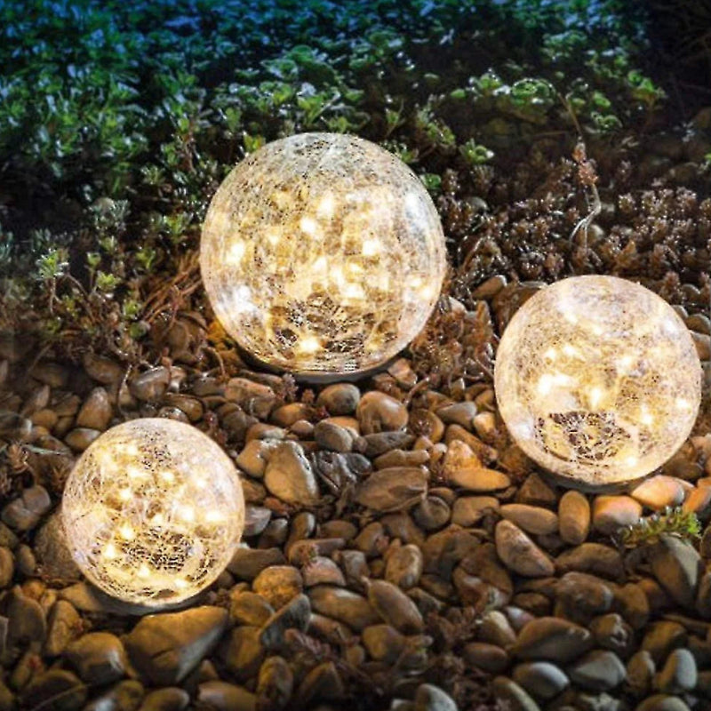 Three solar-powered crackle glass globe lights glowing warmly on a bed of small rocks in an outdoor garden setting at night. The lights create a cozy and decorative ambiance, illuminating the surrounding greenery.