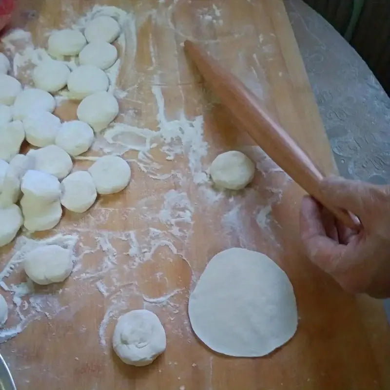 Person using a wooden rolling pin to flatten dough on a floured wooden surface, with small dough pieces scattered around.