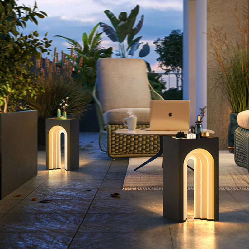 The image shows an outdoor patio scene at dusk. Two modern black side tables with a built-in arched light feature illuminate the area. A comfortable wicker chair sits nearby, and lush greenery surrounds the space, creating a relaxing atmosphere.