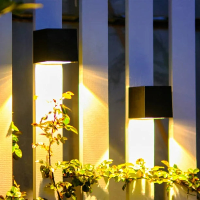 Two modern black cube-shaped wall lights mounted on a white slatted fence, casting warm vertical beams of light. Green climbing plants with small leaves grow along the fence, illuminated by the soft glow of the lights, creating a stylish and inviting outdoor ambiance.