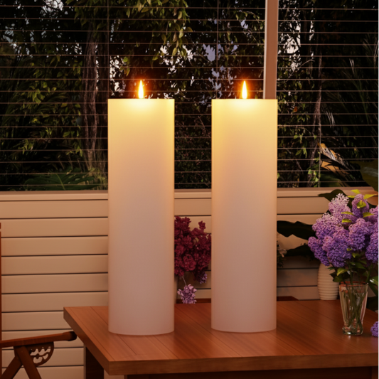 Two tall, glowing white LED pillar candles with faux flames on a wooden patio table at night, with blinds and plants in the background.