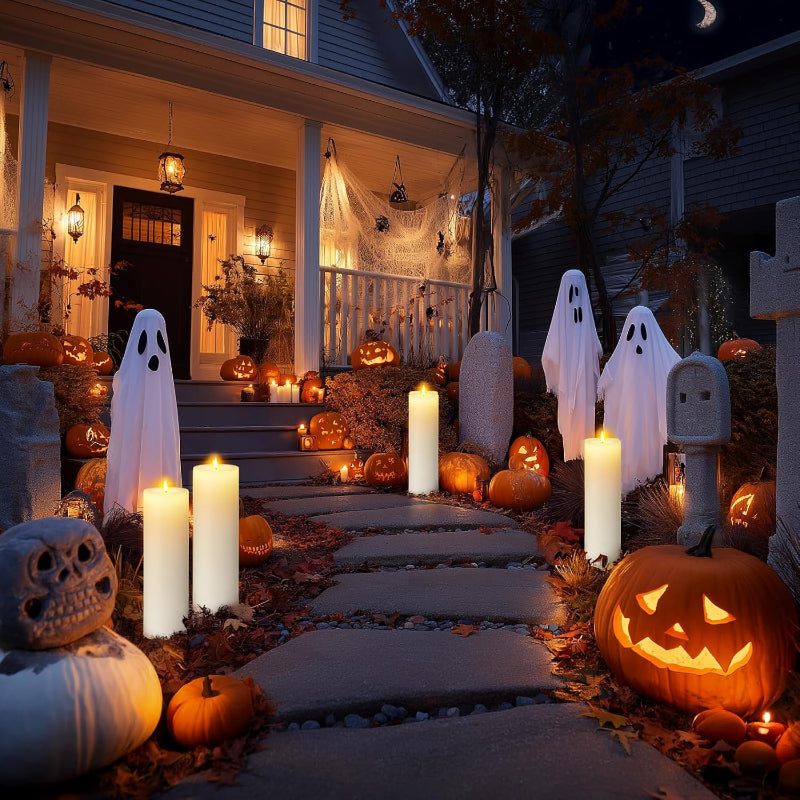 A house porch decorated for Halloween at night with glowing jack-o'-lanterns, sheet ghosts, tombstones, and multiple white LED pillar candles lighting a stone walkway.