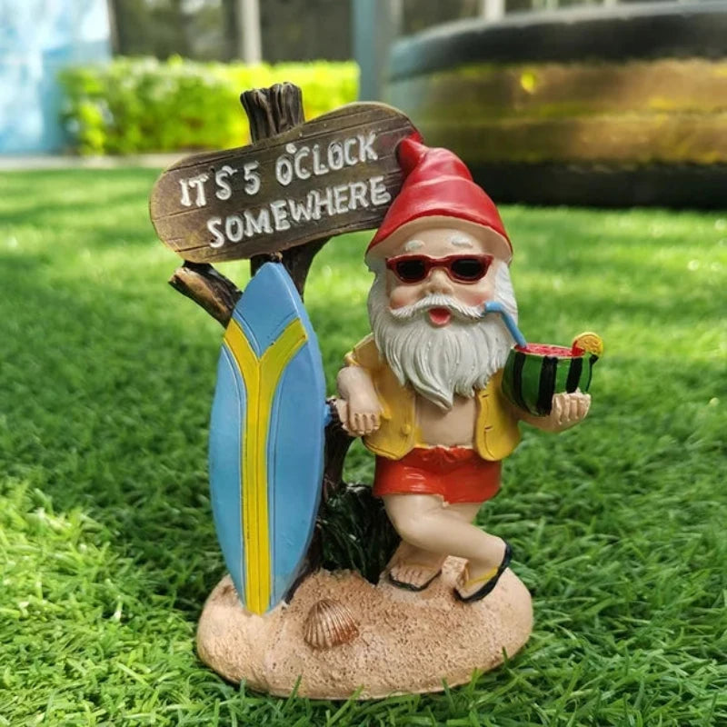 A whimsical garden gnome figurine stands on artificial green grass. The gnome has a long white beard, wears sunglasses, a red pointed hat, a yellow shirt, and red shorts. He holds a slice of watermelon in one hand and a small pipe in the other. Behind him is a rustic wooden sign that reads "IT'S 5 O'CLOCK SOMEWHERE" attached to a small surfboard with blue and yellow stripes. The base of the figurine is sandy with small shells. The background is blurred, showing greenery and a dark object.