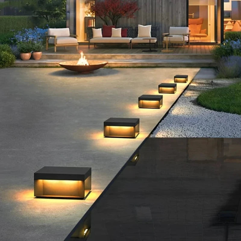 Six modern, square outdoor lights with dark tops and glowing warm-toned panels, illuminating a stone pathway leading to an outdoor living space with a fire pit and seating at dusk.
