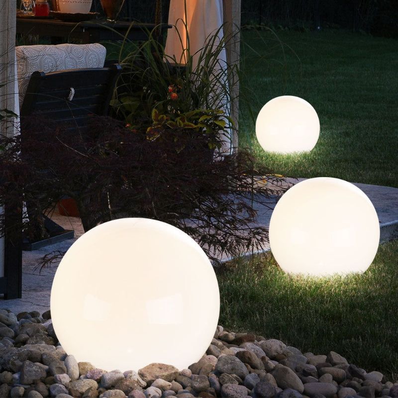 Three glowing white spherical outdoor lamps illuminate a garden area at night. The largest lamp sits on a bed of small rocks, while the other two are on the grass. A glimpse of outdoor furniture and plants is visible in the background.