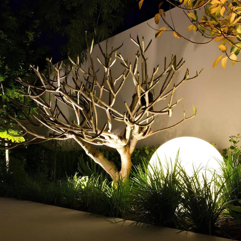 A glowing white spherical lamp illuminates a garden area at night, surrounded by pandanus and lemongrass.