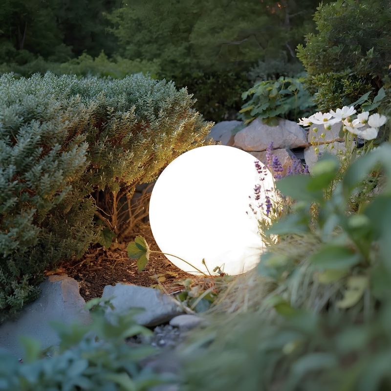 A glowing white spherical outdoor lamp sits among greenery and rocks in a garden at night.