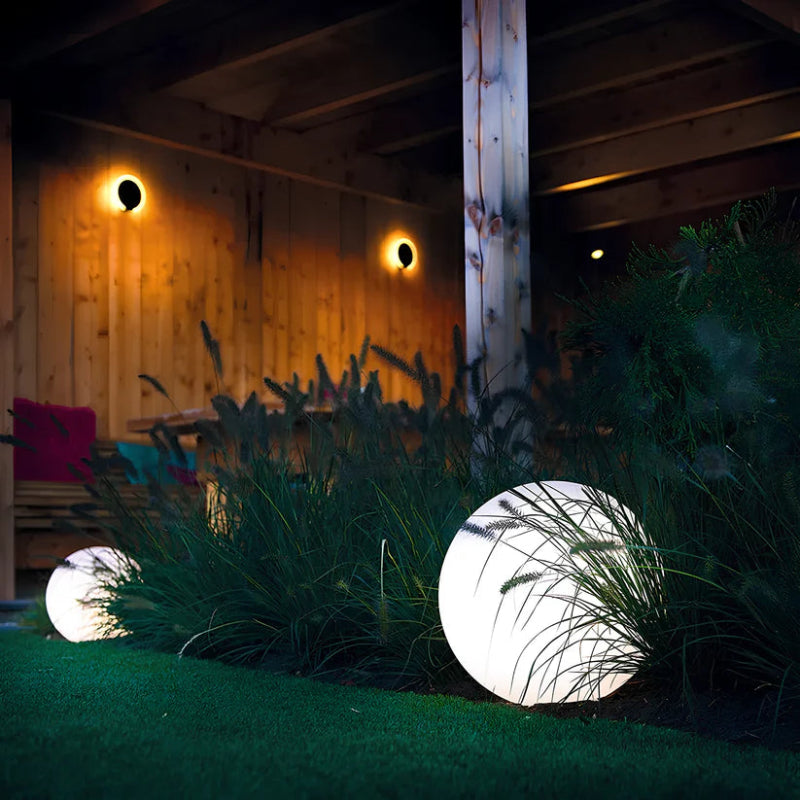 Two glowing white spherical outdoor lamps illuminate a grassy area at night. A larger lamp sits on a bed of small rocks, and a smaller one is on the grass. Some outdoor furniture and plants are visible in the blurred background.