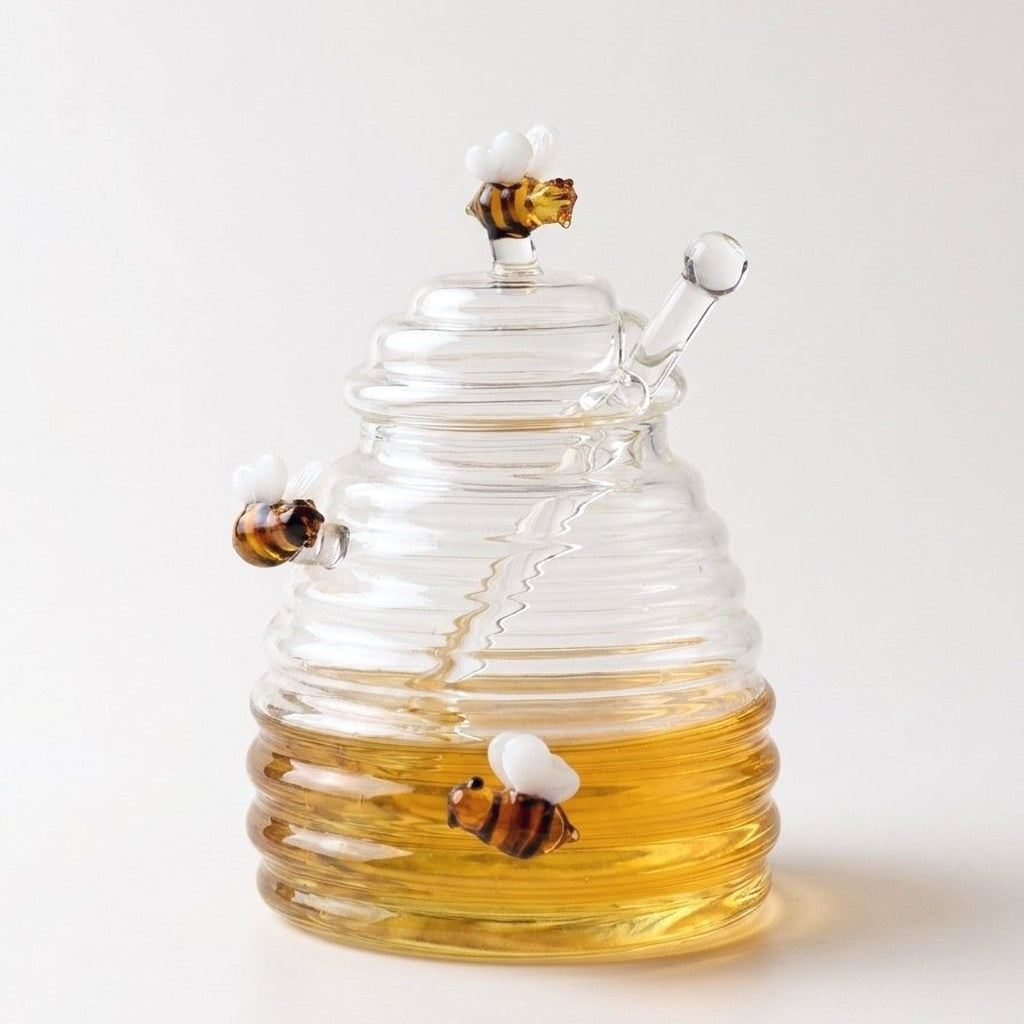 Beehive-shaped glass honey jar with bee accents and dipper on a white background