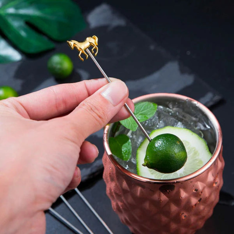 A hand holding a cocktail pick with a gold horse topper, skewering a lime in a copper mug filled with a drink and ice.