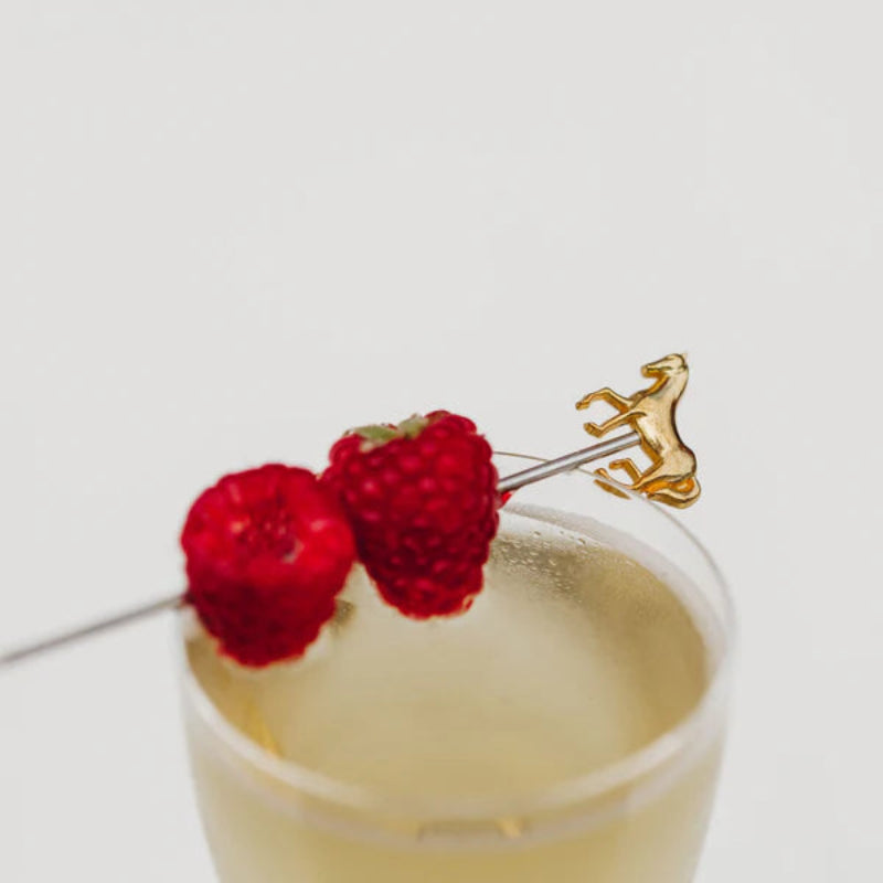 Two raspberries on a cocktail pick with a gold horse topper, in a glass of champagne.