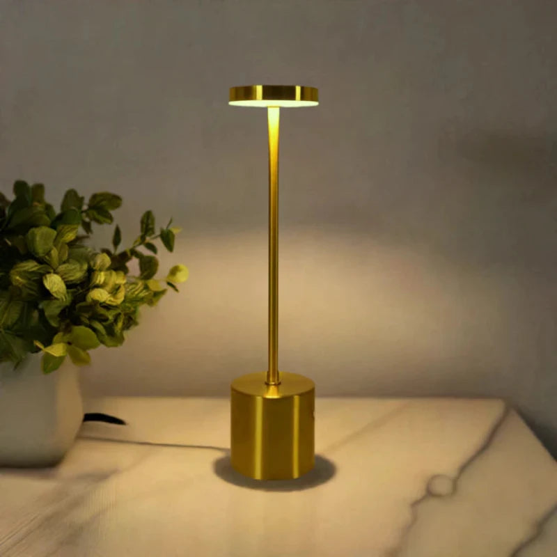 A cordless LED table lamp with a brushed gold finish, featuring a cylindrical base, a slender stem, and a circular light panel emitting a warm downward glow, sitting on a light-colored marble-patterned surface next to a white pot with lush green foliage. The background is dimly lit.