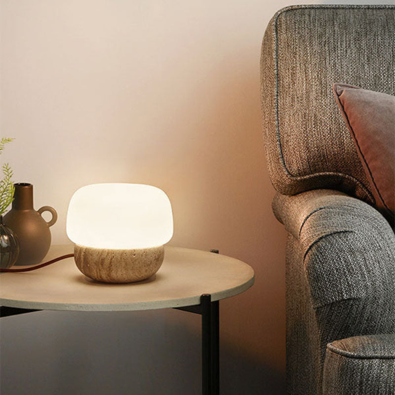 A small, glowing table lamp with a rounded white shade and a textured, light-colored base sits on a round side table with black legs. The lamp's brown cord extends to the left. The table is positioned next to a grey upholstered armchair with a maroon pillow. A dark vase with greenery is partially visible on the table behind the lamp, against a light beige wall.