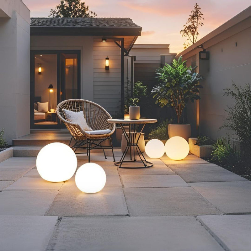 Three glowing white spherical outdoor lamps illuminate a stone-paved patio at dusk. A round table with two chairs, potted plants, and a glimpse into a warmly lit interior space are also visible. The sky shows hues of orange and purple, and a fourth, smaller glowing sphere is visible near the table.