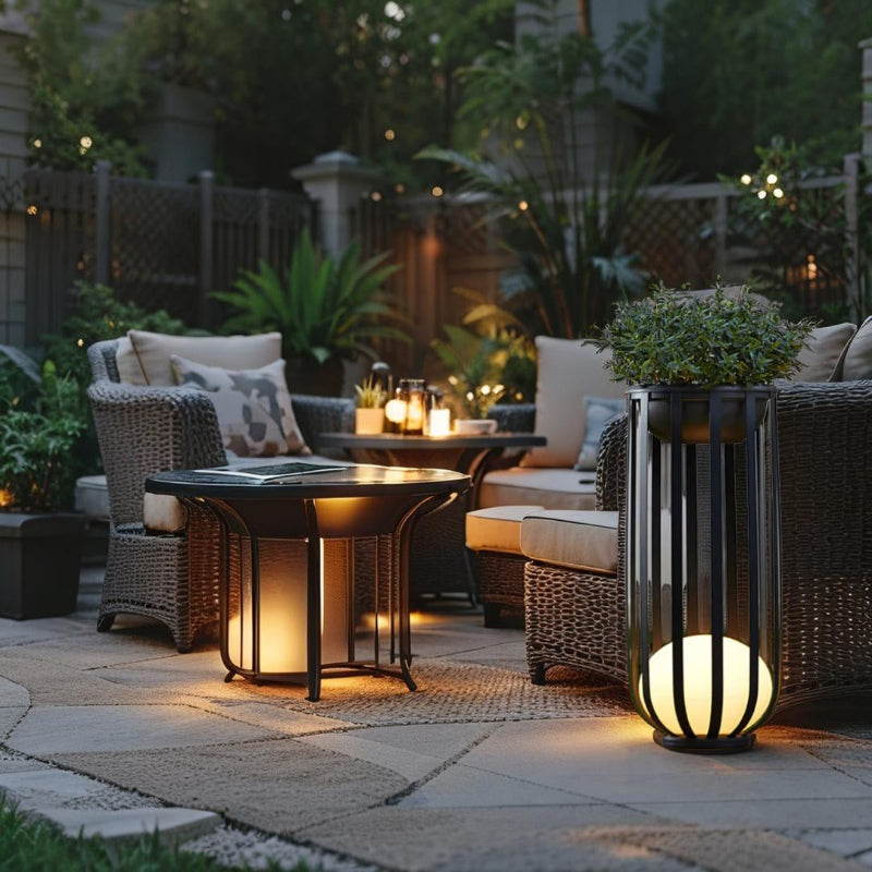 A cozy outdoor patio at dusk features wicker furniture with light cushions arranged around a small round table that emits a warm glow from underneath. A tall black lantern with a glowing white spherical base and a potted plant on top stands nearby. The patio is paved with light-colored stones and decorated with string lights, potted plants, and candles on a separate table in the background, creating a warm and inviting atmosphere.