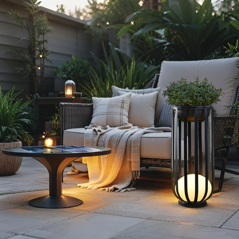 A modern outdoor patio at dusk features a small round black table with a solar panel top, a wicker lounge chair with light cushions and pillows draped with a throw, and a tall black lantern with a glowing white spherical base and a potted plant on top. The patio is paved with light grey tiles and surrounded by lush green plants and a fence. Warm light emanates from the lantern and a smaller lamp on a side table in the background, creating a cozy ambiance