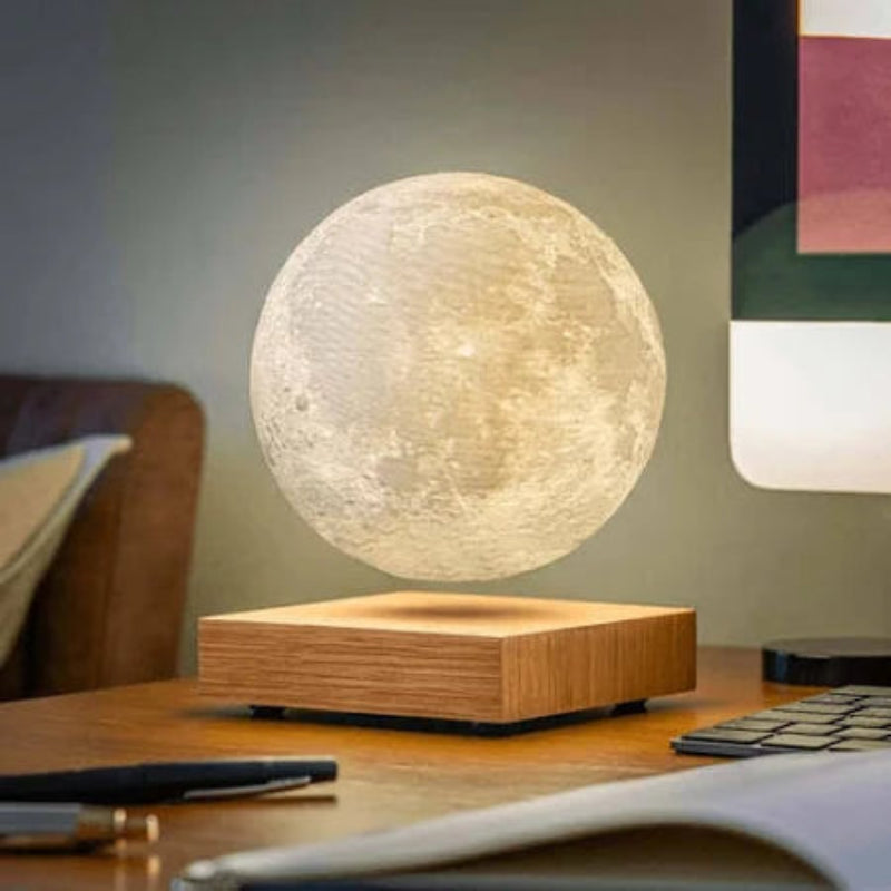 A spherical lamp resembling the moon levitates above a square light wood base that emits a warm upward glow. The lamp's textured surface mimics the moon's craters. The lamp and base sit on a wooden desk. A black pen, a keyboard, and the edge of a computer monitor are also visible on the desk. The background is a muted green-gray wall with a partially visible brown chair.