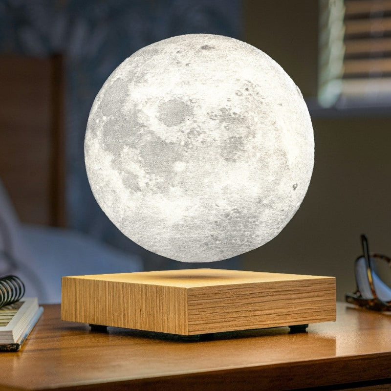 A spherical lamp resembling the moon levitates above a square light wood base that emits a warm upward glow. The lamp's textured surface mimics the moon's craters. The lamp and base sit on a wooden nightstand. A notebook and pen are visible to the left, and eyeglasses are partially visible to the right. The background is softly blurred, suggesting a bedroom setting.