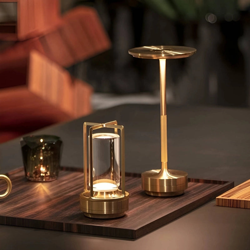 Two cordless LED table lamps with a metallic gold finish sit on a dark wooden tray. The lamp on the right has a slim stem and a disc-shaped shade emitting a warm downward light. The lamp on the left is lantern-shaped with a clear cylindrical diffuser and emits light from the top and bottom. A textured glass candle holder with a flickering light inside is also on the tray. The background is blurred, showing warm-toned architectural details.