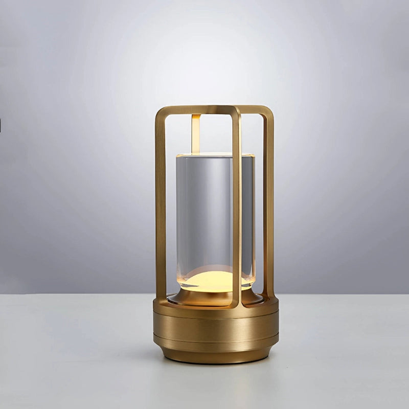 A small, portable LED table lamp with a metallic gold base and frame stands on a white surface against a gradient gray background. The lamp features a frosted cylindrical diffuser that emits a warm glow from the top and bottom. The frame has a simple, open rectangular design