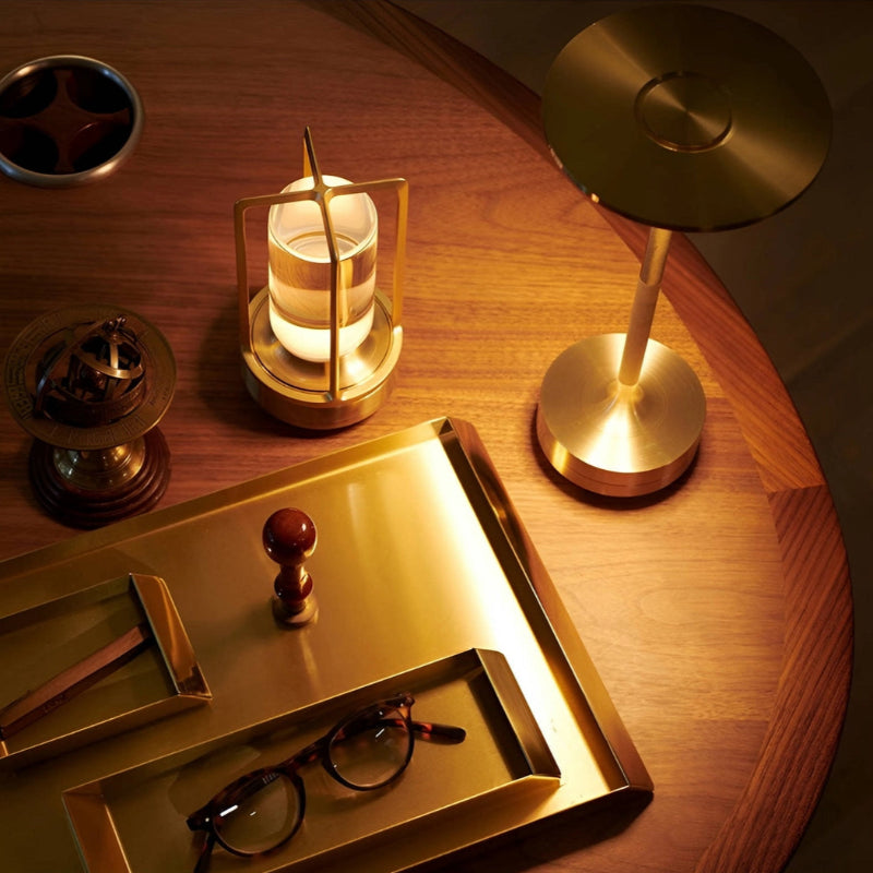Several metallic gold-toned objects are arranged on a rich brown wooden table. These include a cordless table lamp with a disc-shaped shade, a small lantern-style lamp with a frosted glass cylinder, a decorative globe on a stand, a round ashtray, and a rectangular tray holding a pair of round eyeglasses and other small items. The scene is dimly lit, highlighting the metallic sheen of the objects.