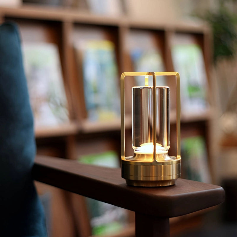 A small, portable LED table lamp with a metallic gold finish and a clear cylindrical diffuser sits on the dark wooden armrest of a chair. The lamp emits a warm glow from its base and top. A blurred background shows a bookshelf filled with books. The armrest is smooth and reflects some light. A portion of a blue velvet chair is visible on the left.