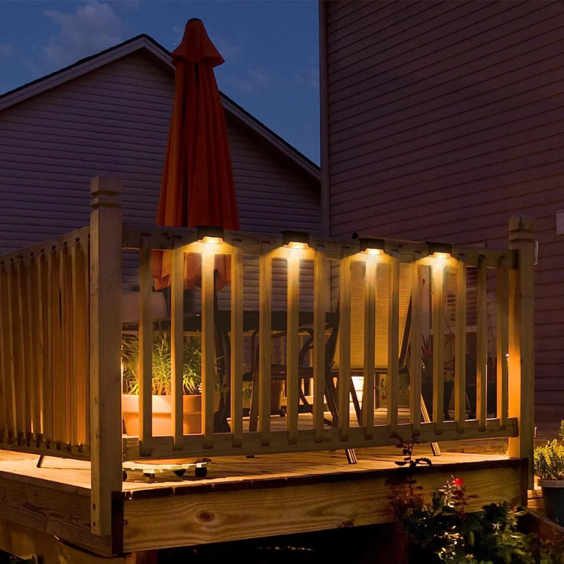 Four small, dark, rectangular solar lights are mounted along the top rail of a wooden deck at dusk, casting a warm yellow downward glow. The deck has wooden railings and appears to have a table and chairs underneath an orange umbrella. The side of a house is visible in the background under a darkening sky.