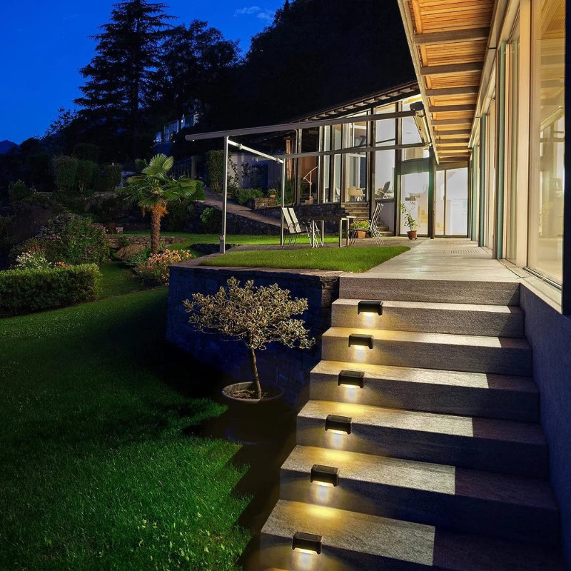 A well-lit outdoor staircase at night features six small, dark, rectangular lights mounted along the sides of the steps, casting a warm white downward glow. Railings flank the stairs, which ascend into the darkness.