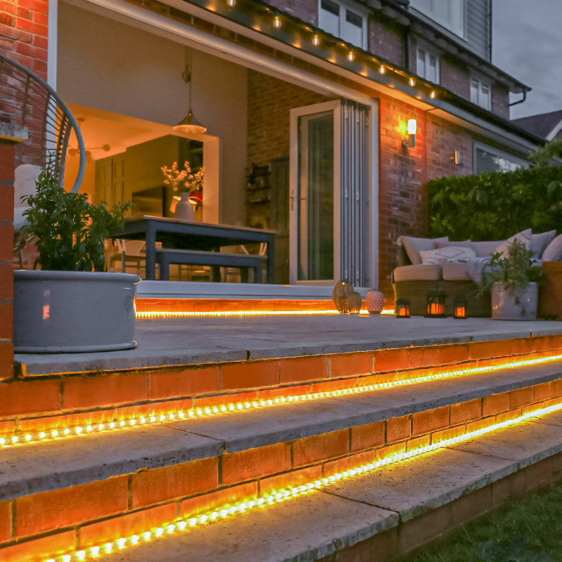 Warm white LED rope lights illuminate the edges of brick and stone outdoor steps leading to a patio at dusk. The patio features a dining table, chairs, a lounge area with cushions, potted plants, and decorative lanterns. String lights hang above, and the interior of the house is softly lit in the background.