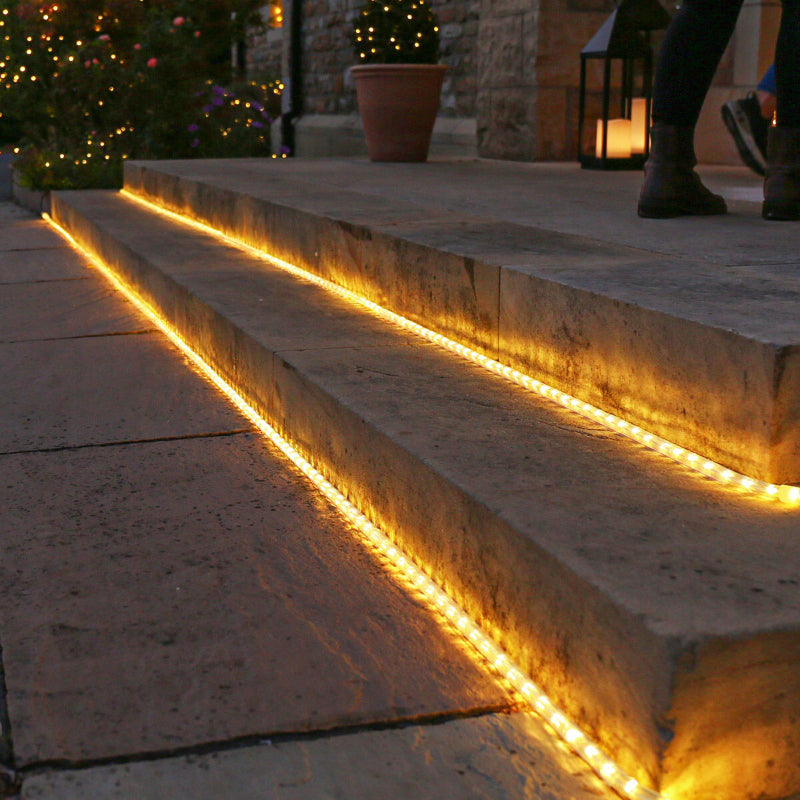 Warm white rope lights illuminate the edges of stone outdoor steps at dusk or dawn. The steps lead upwards, and the feet of a person wearing dark pants and boots are visible at the top. Potted plants and blurred string lights decorate the background, suggesting a festive or cozy outdoor setting.