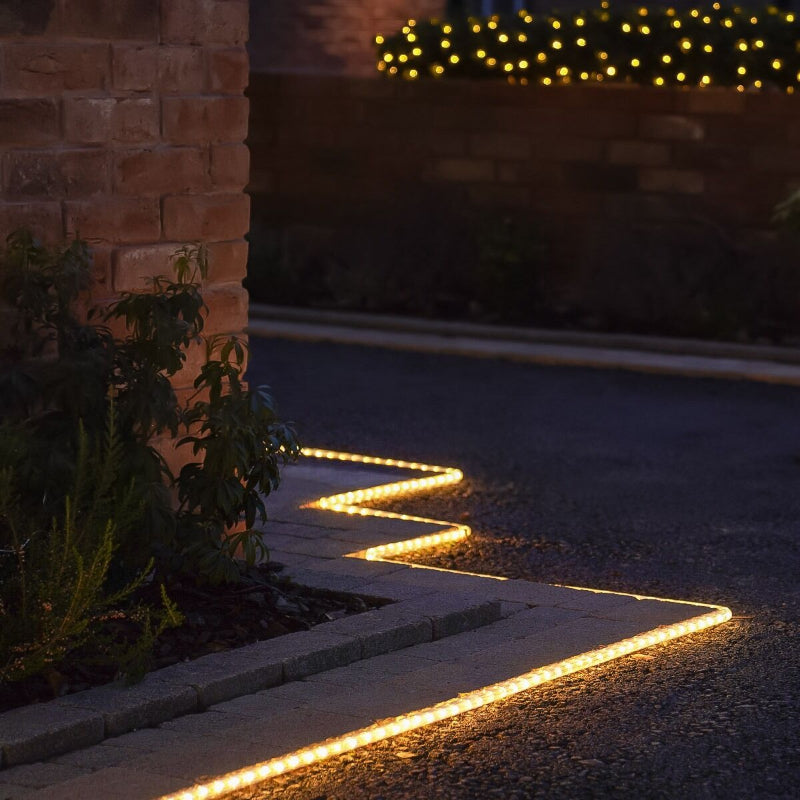 A warm white rope light in a zig-zag pattern illuminates the edge of a dark driveway or walkway at night. The rope light runs alongside a brick wall with some greenery and a border of small paving stones. Soft yellow string lights are visible in the blurred background.