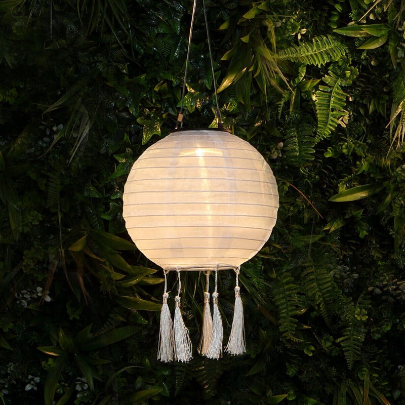 A round, white fabric lantern with tassels hangs illuminated against a backdrop of dark green foliage.