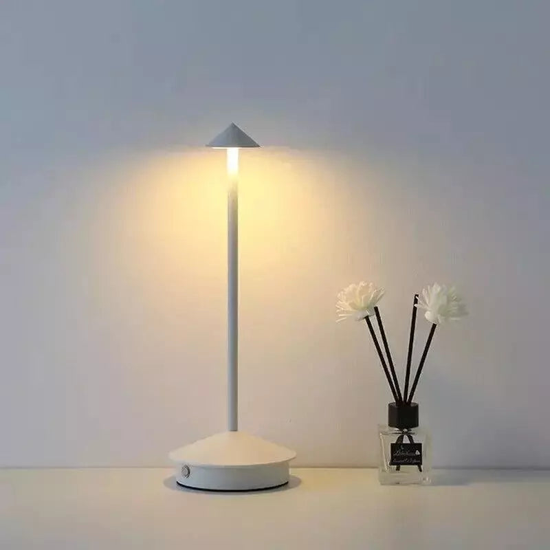 A lit, slender white cordless lamp with a pointed shade stands on a white surface next to a small black diffuser bottle with white artificial flowers. The background is a plain light gray.