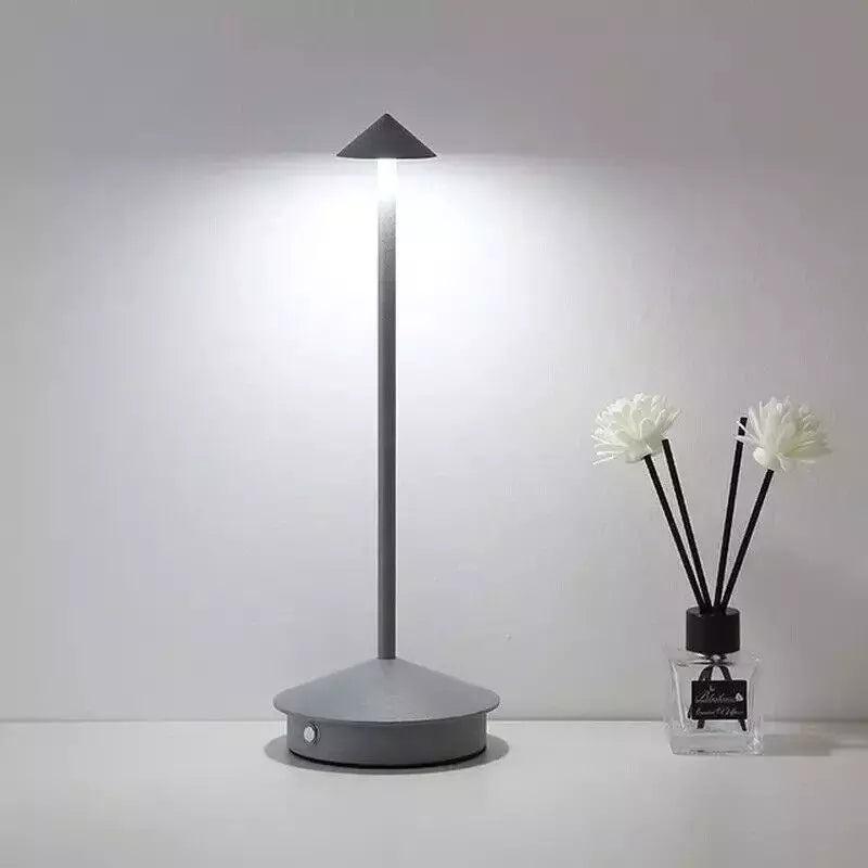 A lit, slender gray cordless lamp with a pointed shade stands on a white surface next to a small diffuser bottle with black reeds and white artificial flowers. The background is a plain light gray.