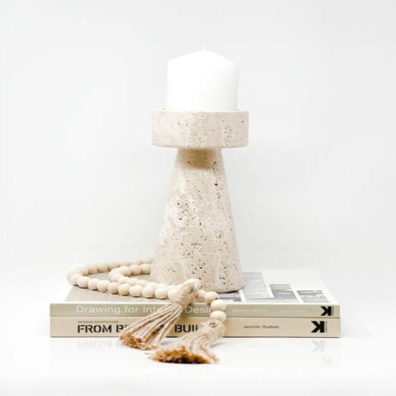 A textured, light-colored faux travertine stone candle holder with a white pillar candle, sitting on a stack of two books, draped with a strand of light wooden beads with tassels, against a bright white background.