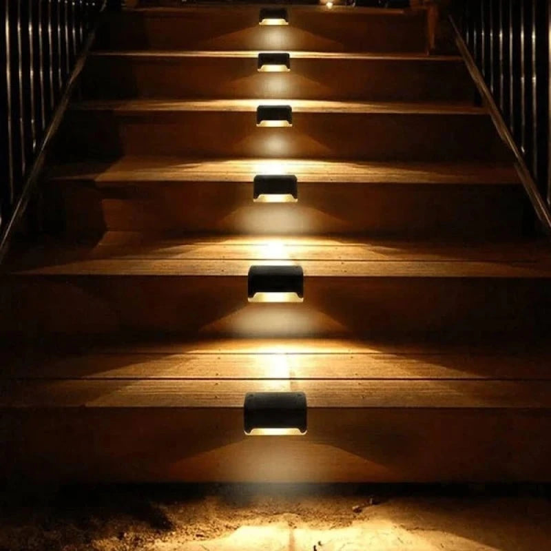 Six small, dark, rectangular solar lights are evenly spaced and mounted along the sides of a set of wooden outdoor stairs at night, emitting a warm white downward glow that illuminates each step. The stairs are flanked by railings and lead upwards. The scene shows illuminated outdoor steps for safety and ambiance.