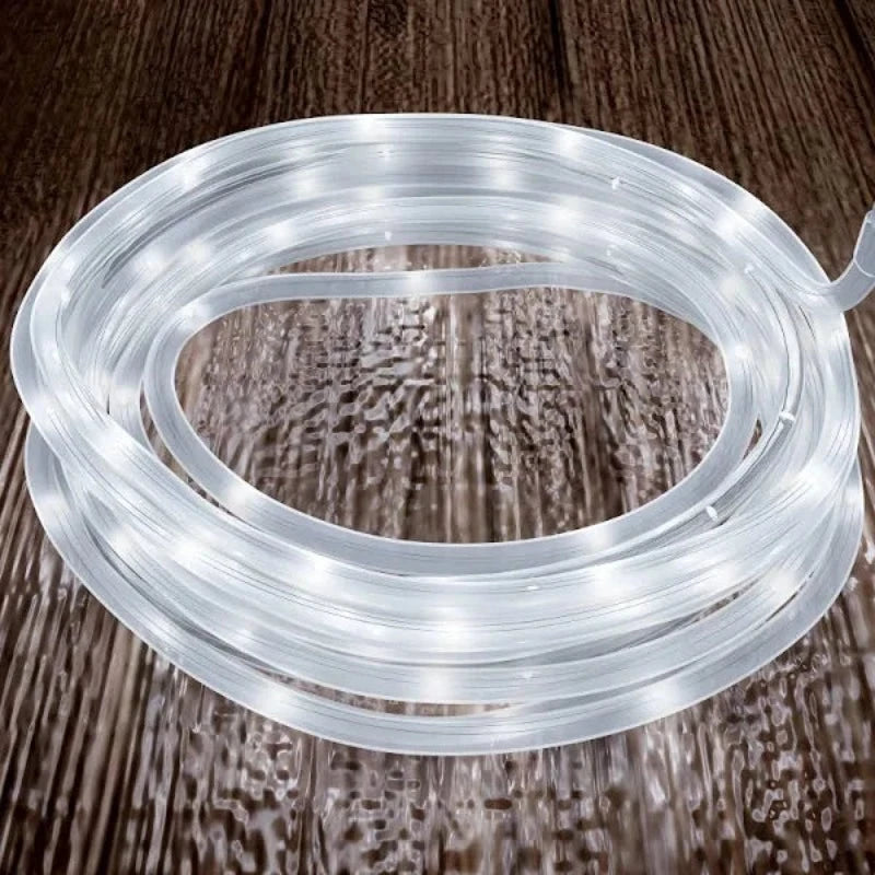 A coiled length of clear rope light emitting a bright white glow sits on a dark brown wooden surface. The rope light is neatly wound in a spiral.
