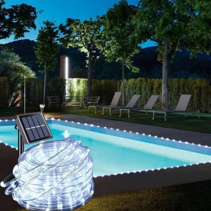 A coiled white solar tube light with an attached solar panel and stake sits on green grass beside a rectangular swimming pool at dusk. The pool's edges are lined with similar white tube lights, illuminating the water. Lounge chairs and trees are visible around the pool under a dark blue sky.